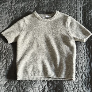 Grå stickad tröja från Zara - Säljer en stilren beige stickad tröja från Zara. Tröjan har korta ärmar och en rund halsringning. Perfekt för lager-på-lager eller som den är. Passar till många olika stilar och tillfällen.