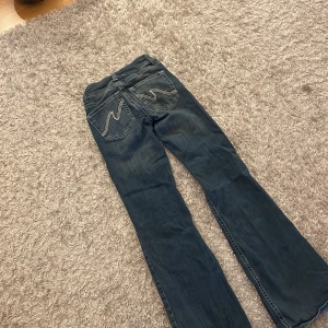 Blå bootcut jeans med broderade bakfickor - Snygga blå bootcut jeans med unika broderade detaljer på bakfickorna. Perfekta för en avslappnad stil med en touch av retro. De har en klassisk femficksdesign och stängs med dragkedja och knapp. 