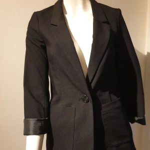 Svart kavaj med knappar - Elegant svart kavaj med klassisk design och knappar framtill. Kavajen har långa ärmar och fickor på sidorna, vilket ger en stilren look. Perfekt för en formell eller professionell stil.