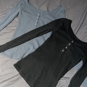 Ribbad långärmad tröja med knappar - Två lite urringade ribbade långärmade tröjor i svart och grå. Båda har en stilren knappdetalj framtill som ger en extra touch. Storlek S/M. 40kronor för båda.