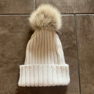 Beige/cremefärgad stickad mössa från Hollies - Snygg stickad mössa från Hollies med en stor fluffig pälsboll på toppen. Pälsbollen är i äkta tvättbjörnspäls.