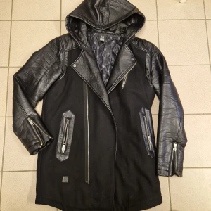 Svart jacka med läderdetaljer från Black Kaviar - Säljer en svart jacka från Black Kaviar med coola läderdetaljer och huva. Jackan har dragkedjor på fickorna och ärmarna, samt en quiltad insida. Materialet är en mix av ull och polyester med syntetiskt läder.