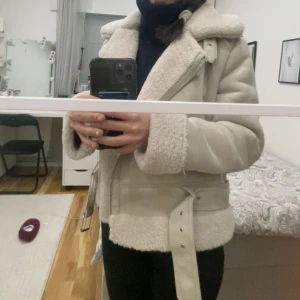 Beige Stradivarius jacka  - Säljer en snygg beige pälsjacka med bälte och dragkedja. Jackan har en mysig krage och långa ärmar med pälsdetaljer. Perfekt för kyliga dagar!