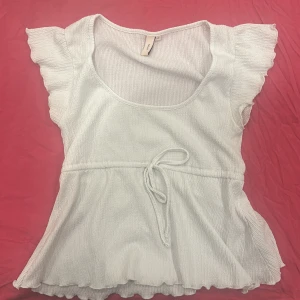 Vit topp från Nelly - Säljer en söt vit topp från Nelly med korta volangärmar och knytband i midjan. Toppen har en rund halsringning och är tillverkad i ett ribbat material som ger en fin struktur. Perfekt för en somrig look!