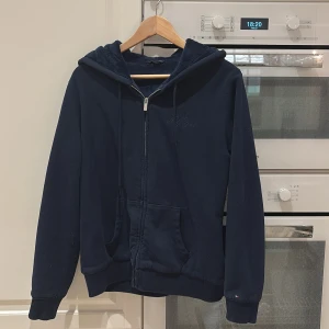 Mörkblå hoodie från Tommy Hilfiger - Säljer en snygg mörkblå hoodie från Tommy Hilfiger med dragkedja och huva. Tröjan har en mjuk insida och ribbade muddar vid ärmslut och nederkant. Perfekt för kyliga dagar! 💙