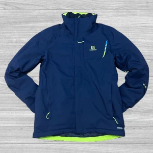 Salomon jacka - Säljer en blå jacka från Salomon med gröna detaljer. Jackan har dragkedja och hög krage, perfekt för kyliga dagar. Den är designad med AdvancedSkin Dry-teknologi för att hålla dig torr och bekväm.