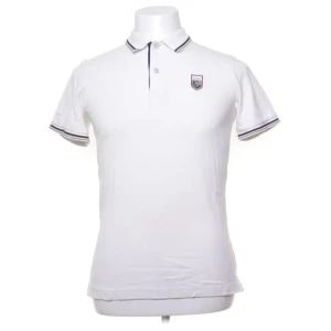 Vit pikétröja från GANT x Le Mans - Säljer en stilren vit pikétröja från GANT x Le Mans. Tröjan har korta ärmar och en klassisk krage med knappar. Den är tillverkad i 100% bomull och har en broderad logotyp på bröstet. Perfekt för en sportig och elegant look.