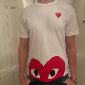 Vit CDG t-shirt - Säljer en vit t-shirt från Comme des Garçons med ett ikoniskt rött hjärtmotiv på bröstet och ett större tryck längst ner. Perfekt för en stilren och trendig look. 