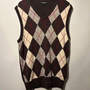 Brun rutig väst från Express Design Studio - Brun stickad väst med ett argylemönster i beige och röda detaljer. V-ringad hals. Bra skick. Har litet hål.