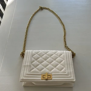 Väska från Gina Tricot - Axelremsväska från Gina Tricot. Créme/vitfärgad. Svart foder. Guldkedja.  Lite avskavd på vridknappen fram men annars i väldigt bra skick.