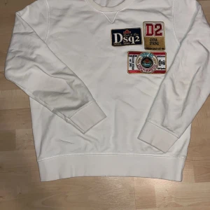 Vit sweatshirt  - Säljer en vit sweatshirt från Dsquared2 med flera broderade märken på framsidan. Tröjan har långa ärmar och en klassisk rund halsringning. Perfekt för en avslappnad stil med en touch av design.