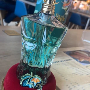 Jean Paul Gaultier Le Beau Paradise Garden - Säljer en unik parfymflaska från Jean Paul Gaultier, Le Beau Paradise Garden. Flaskan har en skulptural design i blågrönt glas med en guldig spraypump. Förpackningen är dekorerad med tropiska mönster i livfulla färger. Perfekt för den som vill ha en doft med karaktär och stil. Det är 80% vätska kvar