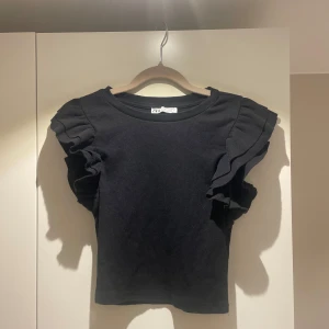 Svart topp med volangärmar från Zara - Säljer en stilren svart topp från Zara med volangdetaljer på ärmarna. Toppen har en ribbad struktur och korta ärmar som ger en trendig look. Perfekt för att piffa upp din garderob!