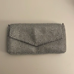 Glittrig silver kuvertväska - Fin kuvertväska i silver med glittriga paljetter. Väskan har en praktisk magnetknapp för enkel öppning och stängning. Endast använd en gång till bal och därav i mycket bra skick. Har även öglor för att sätta på egen kedja om man vill göra det till en väska. För mig har den fått plats med telefon, kort och läppglans 💕