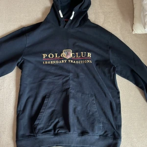 Svart hoodie från Vinson Polo Club - Säljer en svart hoodie från Vinson Polo Club med texten 'Legendary Traditions' på bröstet. Tröjan har en klassisk känguruficka . Perfekt för en avslappnad stil. Har en fläck, kolla sista bilden.