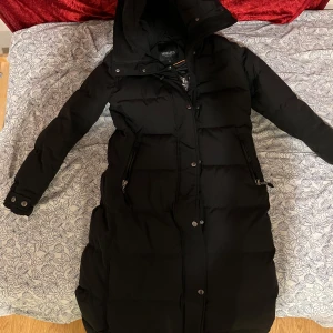 Svart lång dunjacka från Hollies - Säljer en svart lång dunjacka från Hollies. Jackan har en huva och stängs med både dragkedja och knappar. Perfekt för kalla dagar med sin vadderade design.