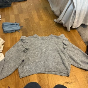 Grå stickad tröja med volangdetaljer - Säljer en mysig grå stickad tröja med volangdetaljer på axlarna. Tröjan har långa ärmar och en rund halsringning. Perfekt för kyliga dagar!