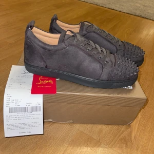 Grå sneakers med nitar från Christian Louboutin - Snygga grå sneakers i mocka från Christian Louboutin med coola nitar på ovansidan. Skorna har den ikoniska röda sulan och snörning för en perfekt passform. Perfekta för att ge din stil en edgy touch. Tar emot trade i storlek 42-43