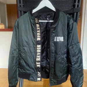Mörk grön bomberjacka från Forever 21 - Snygg mörk grön bomberjacka från Forever 21 med texten 'AU REVOIR' på bröstet och insidan av dragkedjan. Jackan har en dragkedja framtill och en extra ficka med dragkedja på ärmen. Även två vanliga jackfickor. Perfekt för en cool och avslappnad stil.