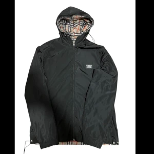 Svart burberry vindjacka  - Snygg burberry vindjacka med ett klassiskt rutigt mönster på insidan. Jackan har dragkedja och justerbara snören vid fållen för bästa passform. Perfekt för blåsiga dagar! Både sidig! DET STÅR XXL MEN DEN PASSAR ÄVEN L