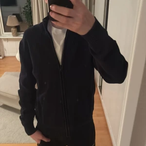 Svart kofta från Dressmann - Säljer en stilren mörk blå kofta från Dressmann med dragkedja framtill. Koftan har en ribbad krage och långa ärmar, perfekt för kyligare dagar. Passar bra till både jeans och chinos.