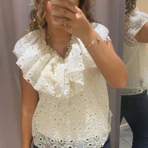 Vit blus från Wera - Charmig vit blus från Wera med broderade detaljer och volangkrage. Perfekt för en romantisk look med sitt vackra mönster och luftiga design. Helt slutsåld!