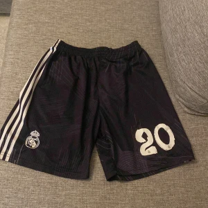 real madrid shorts - Snygga svarta fotbollsshorts med vita ränder på sidorna och nummer 20 tryckt på framsidan. Perfekta för fotbollsträningen eller matchen. Elastisk midja för bekväm passform.