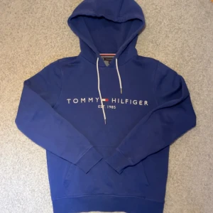 Blå hoodie från Tommy Hilfiger - Säljer en snygg blå hoodie från Tommy Hilfiger med vit logotyp på bröstet. Den har en klassisk design med justerbar huva och långa ärmar. Perfekt för en avslappnad stil. Storlek XS, kan även passa S.