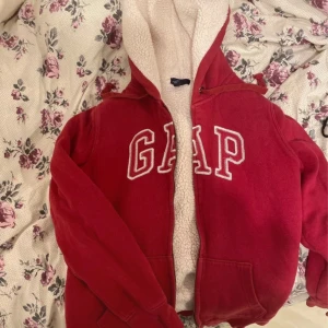 Röd hoodie fluffig från GAP - Säljer en mysig röd hoodie från GAP med vit teddyfoder inuti. Den har en dragkedja framtill och stora bokstäver på bröstet. Perfekt för kyliga dagar och nu till vintern!