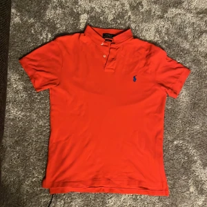 Röd kortärmad pikétröja från Ralph Lauren - Säljer en snygg röd pikétröja från Ralph Lauren med klassisk krage och knappar. Den har en broderad blå logga på bröstet och är i slim fit. Perfekt för en sommar kväll. Säljer på grund utav att den har blivit förliten för mig!