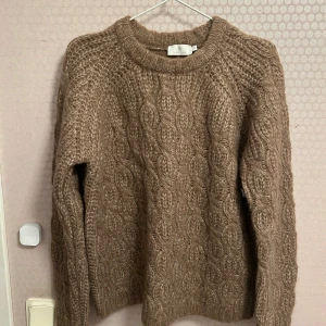 Beige stickad tröja från B. Coastline - Säljer en mysig beige/ brun  stickad tröja från B.coastline . Tröjan har ett klassiskt flätmönster och är tillverkad i en blandning av återvunnen polyester, akryl och ull. Perfekt för kyliga dagar med sin långärmade design. Aldrig använd men ingen prislapp kvar.
