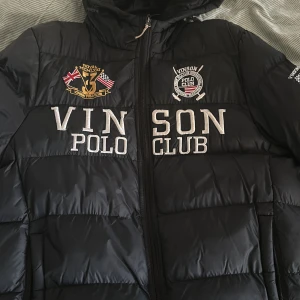 Svart dunjacka | Vinson Polo Club - Snygg svart dunjacka från Vinson Polo Club med broderade detaljer och flaggor. Jackan har en dragkedja framtill och en huva för extra värme. Perfekt för kyliga dagar. Pris går att diskuteras!