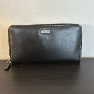 Svart plånbok från Calvin Klein - Snygg svart plånbok från Calvin Klein med diskret logotypmönster och dragkedja runtom. Plånboken har flera fack för kort och en innerficka med dragkedja för mynt. Perfekt för att hålla ordning på dina tillhörigheter med stil.