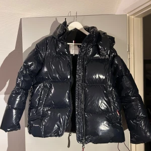 Svart pufferjacka från Tommy Hilfiger - Säljer en svart pufferjacka från Tommy Hilfiger med glansig yta och huva. Jackan har dragkedja framtill och är perfekt för kyligare dagar. Den är stilren och enkel att matcha med olika outfits. Helt ny.
