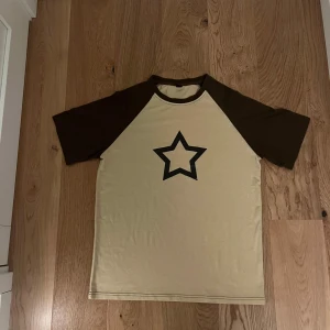 Beige och brun t-shirt med stjärna från Romwe - Säljer en beige t-shirt från Romwe med bruna ärmar och en stor mörkbrun stjärna på framsidan. Perfekt för en avslappnad stil. T-shirten har korta ärmar och en rund halsringning.