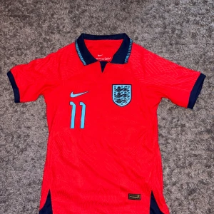 Player Version England Vm 2022 tröja med Rashford - Player Version! Säljer en röd fotbollströja från Nike med nummer 11 och Rashford på baksidan. Tröjan har korta ärmar och englands emblem på bröstet. Perfekt för fotbollsfans!
