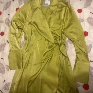 Grön omlottklänning från H&M - Snygg grön omlottklänning från H&M i ett glansigt material. Klänningen har långa ärmar och en knytning i midjan för en smickrande passform. Perfekt för en stilren look.