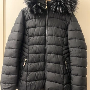 Svart dunjacka från Polo Club - Snygg svart dunjacka från Polo Club med pälskantad huva. Jackan har en quiltad design och dragkedja framtill. Perfekt för kyliga dagar med sin varma och stilrena look.