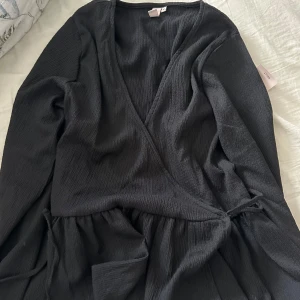 Svart omlott tröja  - Snygg svart omlott tröja  med v-ringning och knytning i midjan. Tröja  har långa ärmar och en elegant, ribbad textur. Den är helt ny och har aldrig används.