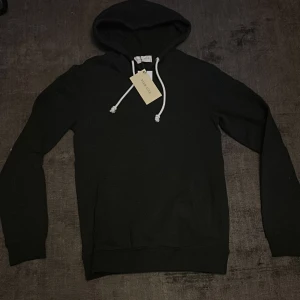 Svart hoodie från Pier One - Säljer en helt ny svart hoodie från Pier One som är helt oanvänd. Den har en klassisk design med justerbara snören i huvan och ribbade muddar vid ärmslut och nederkant. Perfekt för en avslappnad look. Skick 10/10