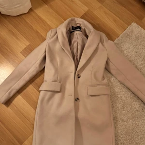 Beige kappa från Boohoo - Snygg beige kappa från new yorker med klassisk design och knappar framtill. Den har en elegant krage och långa ärmar, perfekt för kyligare dagar. Passar bra till både vardag och mer uppklädda tillfällen. Använd någon gång 