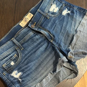 Blå jeansshorts från Hollister - Snygga blå jeansshorts från Hollister med slitna detaljer och fransiga kanter. De är lågmidjade och har klassiska femficksdesign. Perfekta för en avslappnad stil.