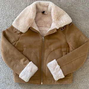 Mocha jacka med luddiga muddar - Ljusbrun mocha jacka med vita luddiga muddar. Luddig på hela insidan och därför varm. Storlek:36