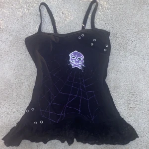 Svart linne med dödskallemotiv - Säljer ett coolt svart linne med smala axelband och ett lila dödskallemotiv på framsidan. Linnet har ett spindelnätsmönster i lila och är dekorerat med nitar och säkerhetsnålar. Nederkanten har en snygg spetsdetalj som ger en extra touch. 