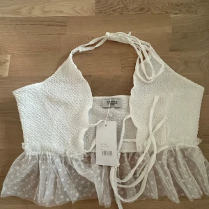 Vit topp med spetsdetaljer från Chiquelle - Säljer en superfin vit topp från Chiquelle i storlek Large. Den har en ribbad struktur och knytband vid axlarna. Toppen är croppad och har en söt spetsdetalj i nederkant med prickigt mönster. Perfekt för sommarens fester eller en dag på stan!