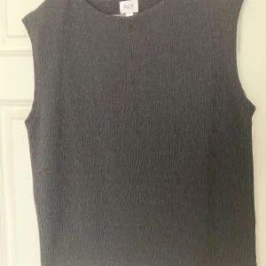 Glittrig ärmlös topp från Saint Tropez - Snygg glittrig ärmlös topp från Saint Tropez i storlek S. Perfekt för festliga tillfällen med sitt stilrena och eleganta utseende. Toppen är i en mörkgrå nyans med en subtil glittrig effekt som ger en extra touch av glamour. Passar perfekt till både kjol och byxor!