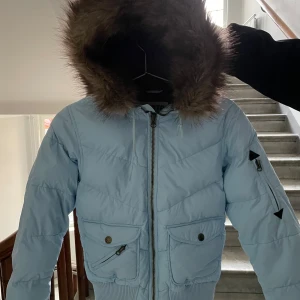 Ljusblå vinterjacka  - En ljusblå jacka med mörkbrun pälshuva i imitation som är avtagbar. Jackan har två fickor framtill och en ficka på vänster ärm. Dunfri och varm