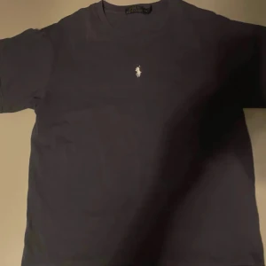 Mörkblå t-shirt från Ralph Lauren - Säljer en snygg mörkblå t-shirt från Ralph Lauren i bomull. Den har en klassisk passform och korta ärmar, perfekt för vardagsbruk. Den ikoniska loggan är broderad på bröstet i vitt. Passar både till jeans och shorts!