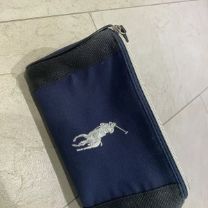 Blå polo Ralph louren necessär  - Säljer en praktisk blå necessär med dragkedja. Den har en stilren design med en broderad logga på framsidan. Perfekt för att hålla ordning på dina småsaker när du är på språng. Materialet verkar vara slitstarkt och lätt att rengöra. Perfekt för resor eller dagligt bruk!