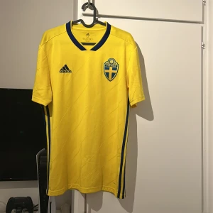 Svenska landslags tröja hemma 18/19.        (PRIS KAN DISKUTERAS) - Säljer en svensk landslags fotbollströja från Adidas med Sveriges landslagsemblem på bröstet. Tröjan har korta ärmar och en blå randig detalj på sidorna. Perfekt för fotbollsfans som vill visa sitt stöd för Sverige! 🟡🔵.                                       Nuvarande pris i SVFF shopen är 749kr